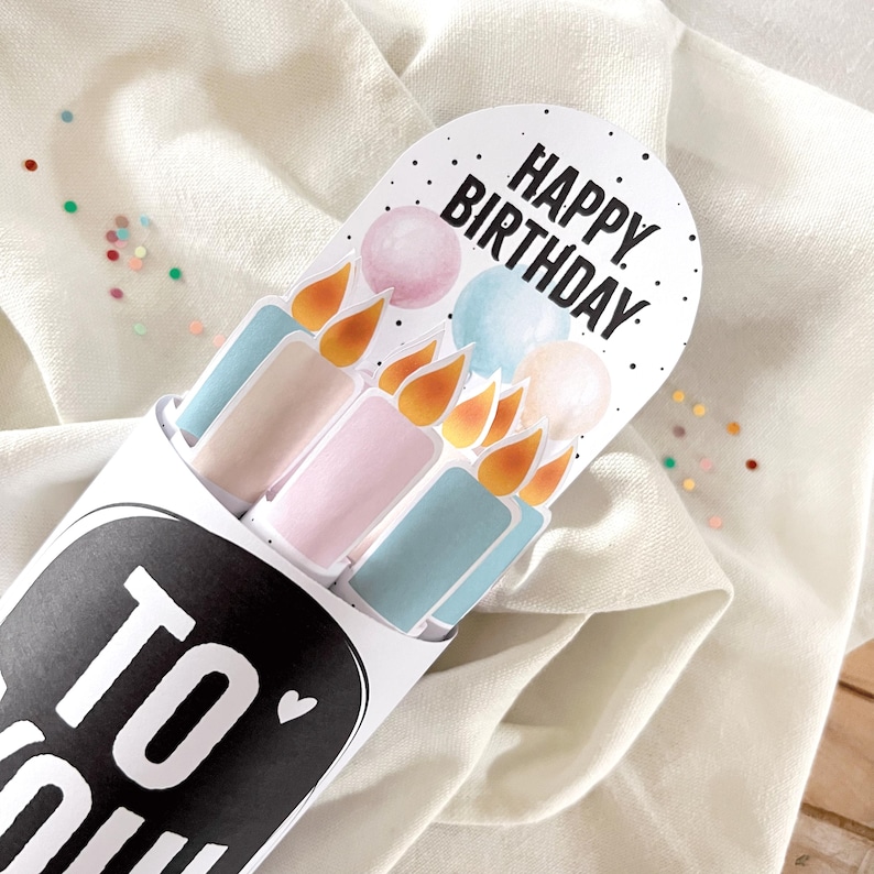 May include: A white paper tube with the words "TO YOU" in black. Inside are pastel-colored candle-shaped cards with the words "HAPPY BIRTHDAY" on top. The candles have orange flames and are pink, blue, and beige.