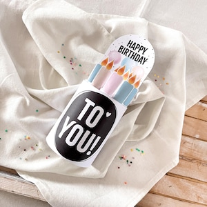 May include: A white birthday card holder with the text "TO YOU!" in a black oval. The card features illustrations of candles in pastel colors and the words "HAPPY BIRTHDAY". The card is on a white cloth with colorful confetti.