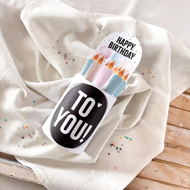 May include: A white birthday card holder with the text "TO YOU!" in a black oval. The card features illustrations of candles in pastel colors and the words "HAPPY BIRTHDAY". The card is on a white cloth with colorful confetti.
