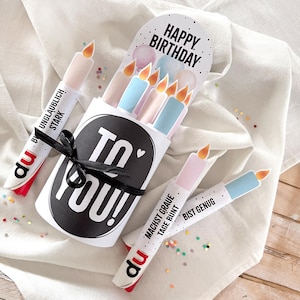 May include: A birthday card set featuring candle-shaped cards with the words "To You!" and "Happy Birthday." The candles are in pastel colors with orange flames. Some cards have German text. Tied with a black ribbon.