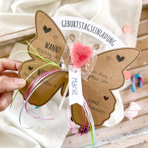 May include: A brown butterfly-shaped card with a pink heart lollipop attached to it. The card has the text "Birthday Invitation" and "When? Friday, the ... Where? Musterra&szlig;e 7" written on it. The card also has a white tag with the name "Marie" written on it.