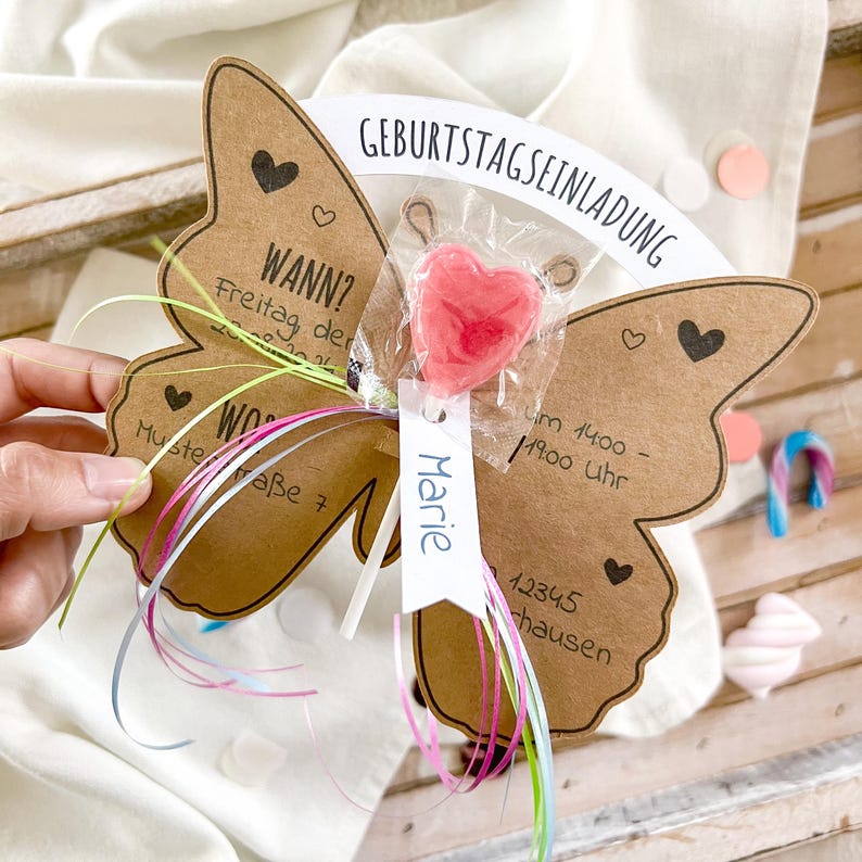 May include: A brown butterfly-shaped card with a pink heart lollipop attached to it. The card has the text "Birthday Invitation" and "When? Friday, the ... Where? Musterra&szlig;e 7" written on it. The card also has a white tag with the name "Marie" written on it.