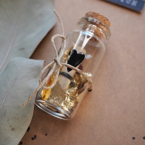May include: A small glass bottle with a cork top is filled with gold leaf and a rolled up piece of paper. A black heart charm is tied to the bottle with twine.