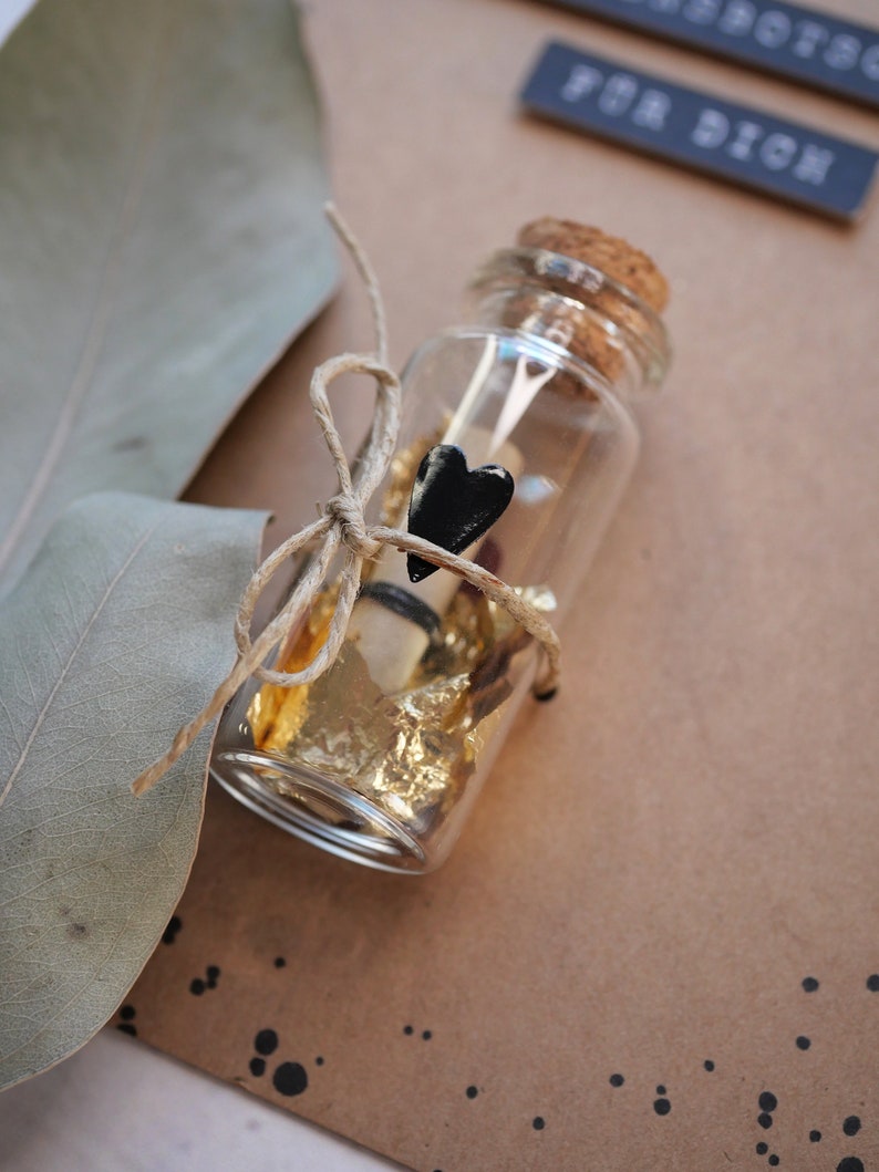 May include: A small glass bottle with a cork top is filled with gold leaf and a rolled up piece of paper. A black heart charm is tied to the bottle with twine.