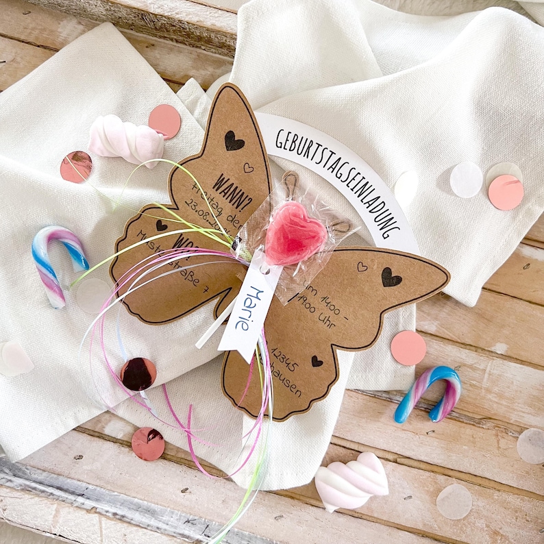 May include: A brown butterfly-shaped card with a pink heart lollipop attached. The card has the text "GEBURTSTAGSEINLADUNG" at the top and "WANN? Freitag der 23.08.20 im 1400 - 1900 Uhr Muststra&szlig;e 7 Marie 12345 hausen" on the bottom.