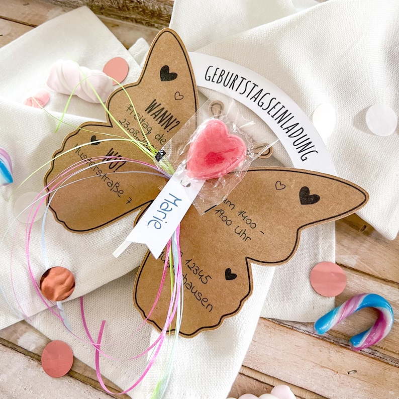 May include: A brown butterfly-shaped card with the text "Geburtstagseinladung" and "Wann? Freitag, de 23.08.2023" and "WO? Musterstra&szlig;e 7" and "Marie" and "um 14:00 - 17:00 Uhr" and "12345 Hausen". A pink heart-shaped lollipop is attached to the card with a string.