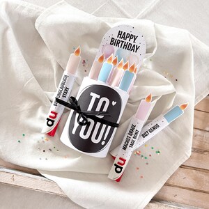 May include: A birthday-themed gift set featuring a white box with the text "TO YOU!" and a black ribbon. The set includes candle-shaped items with the words "HAPPY BIRTHDAY" and other German text. The candles are in pastel colors.
