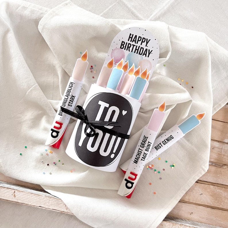 May include: A birthday-themed gift set featuring a white box with the text "TO YOU!" and a black ribbon. The set includes candle-shaped items with the words "HAPPY BIRTHDAY" and other German text. The candles are in pastel colors.
