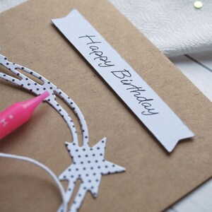 May include: A brown card with a white banner that says "Happy Birthday" in black cursive writing. The card has a white star with black polka dots on it.