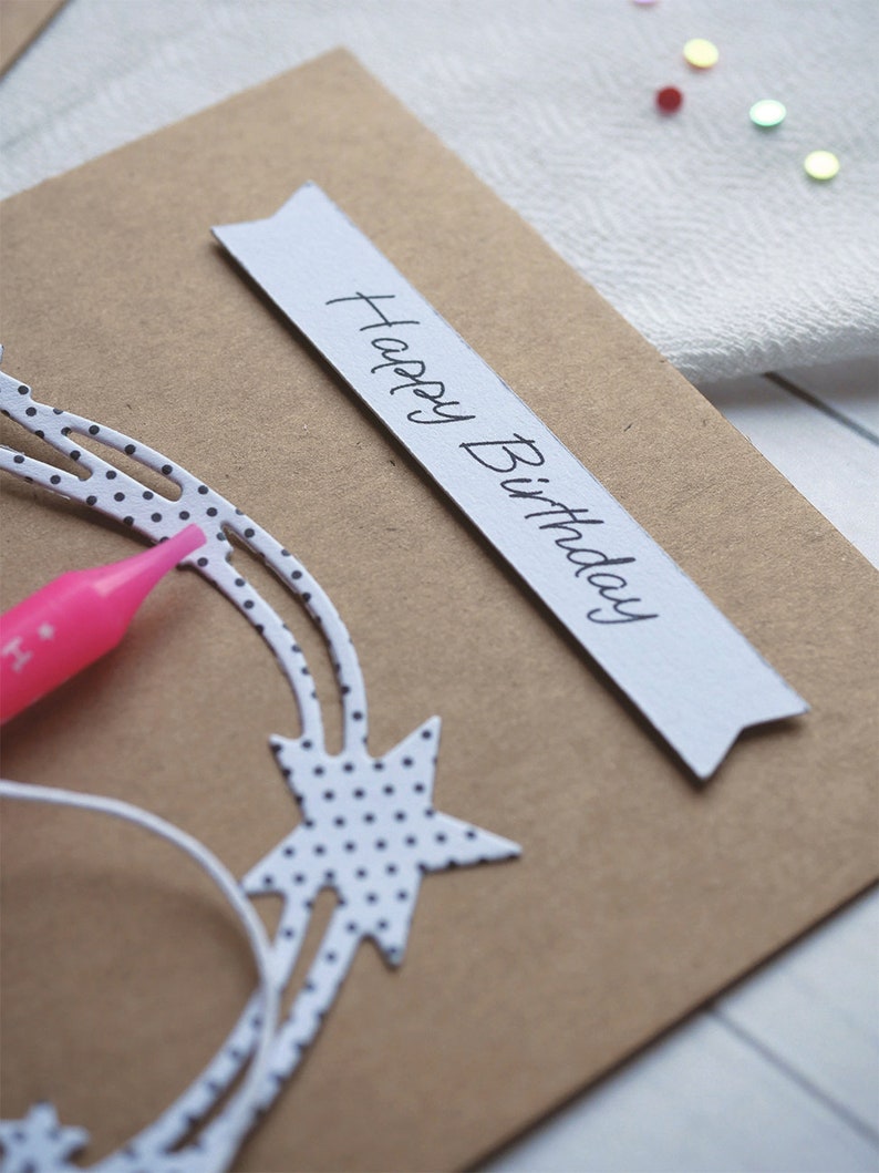May include: A brown card with a white banner that says "Happy Birthday" in black cursive writing. The card has a white star with black polka dots on it.