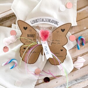 May include: A brown butterfly-shaped card with a pink heart lollipop attached. The card has the text "Geburtstagseinladung" at the top, and the text "WANN? Freitag der 23.08.20 Wo Muststra&szlig;e 7 Marie um 1400 - 1700 Uhr 12345 hausen" on the bottom.