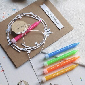 May include: A brown card with the words "Happy Birthday" written on it. The card has a white paper cutout of a star with black polka dots. A pink birthday candle is attached to the cutout. Four additional birthday candles in blue, green, orange, and yellow are on a white wooden surface.