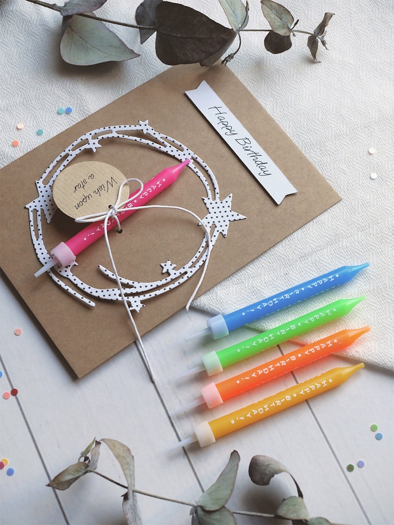 May include: A brown card with the words "Happy Birthday" written on it. The card has a white paper cutout of a star with black polka dots. A pink birthday candle is attached to the cutout. Four additional birthday candles in blue, green, orange, and yellow are on a white wooden surface.