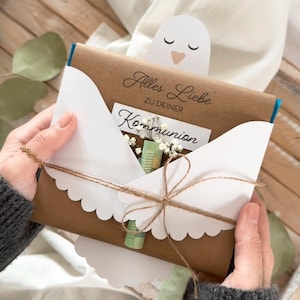 Può includere: Una busta di carta marrone con bordi smerlati bianchi, legata con spago, contenente una carta con le parole "Alles Liebe" e "Kommunion". Una piccola carta bianca a forma di uccello spunta dalla parte superiore. La busta contiene un piccolo bouquet di fiori.