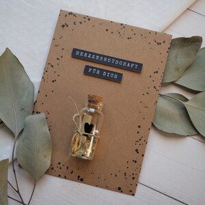 May include: A brown card with black speckled paint. The card has two black labels that read "Herzensbotschaft" and "F&uuml;r Dich". A small glass bottle with a cork top is tied with twine and contains gold glitter and a black heart.