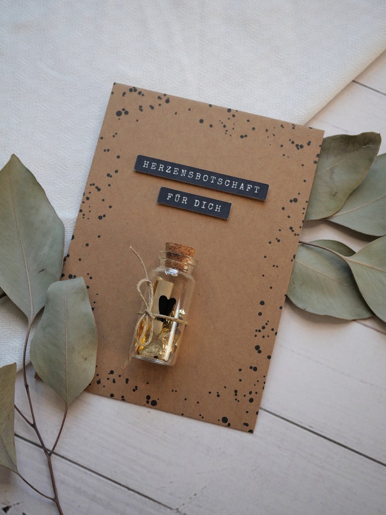 May include: A brown card with black speckled paint. The card has two black labels that read "Herzensbotschaft" and "F&uuml;r Dich". A small glass bottle with a cork top is tied with twine and contains gold glitter and a black heart.