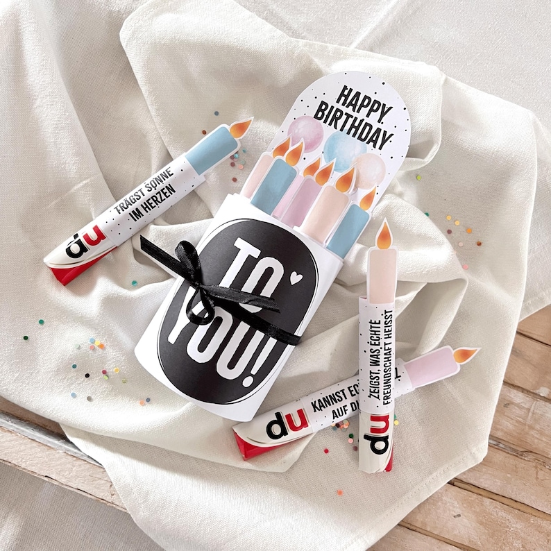 May include: A birthday-themed gift set featuring candle-shaped paper tubes with text and a black ribbon. The set includes a card that says "Happy Birthday" and candles in pastel colors. The tubes have German text.