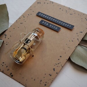 May include: A brown card with black text that reads "Herzensbotschaft" and "F&uuml;r Dich". A small glass bottle with a cork stopper is tied with twine and contains a rolled up piece of paper and gold flakes. The bottle is on a brown background with black dots.