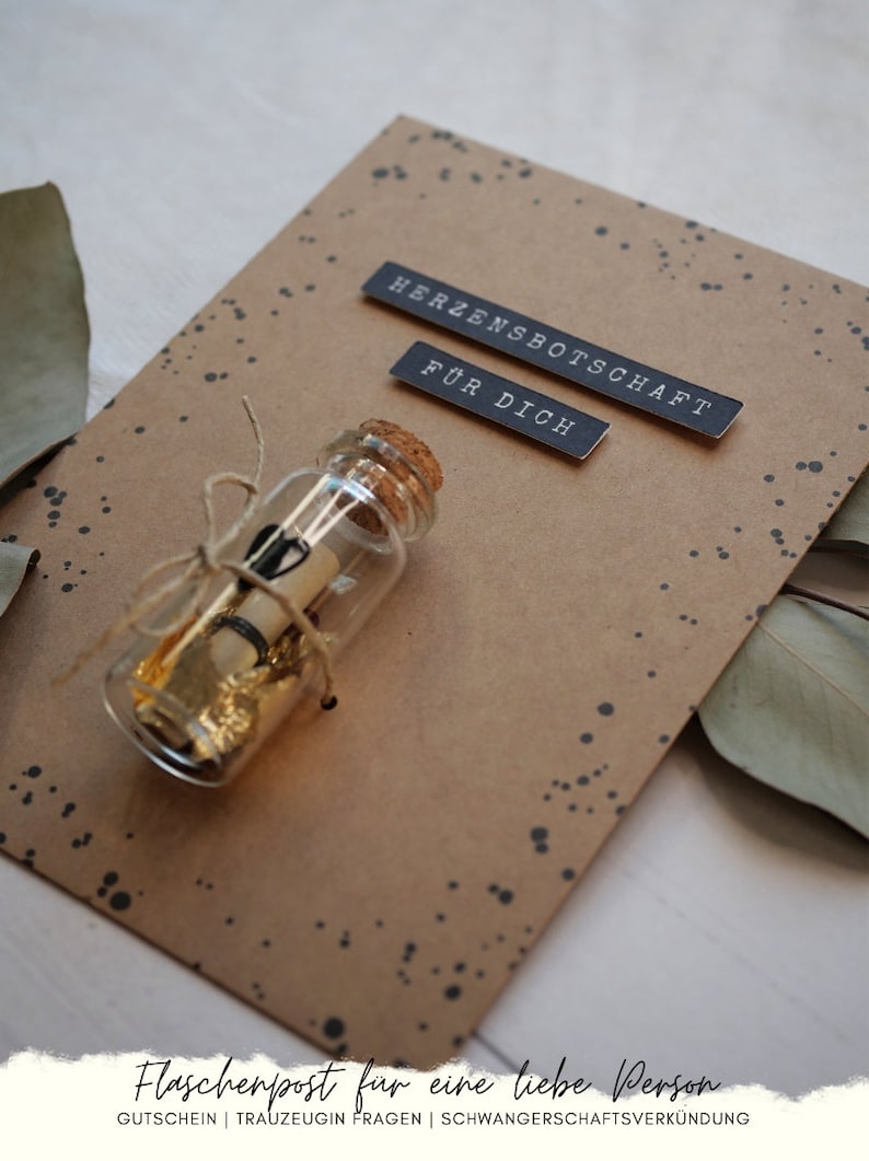 May include: A brown card with black text that reads "Herzensbotschaft" and "F&uuml;r Dich". A small glass bottle with a cork stopper is tied with twine and contains a rolled up piece of paper and gold flakes. The bottle is on a brown background with black dots.