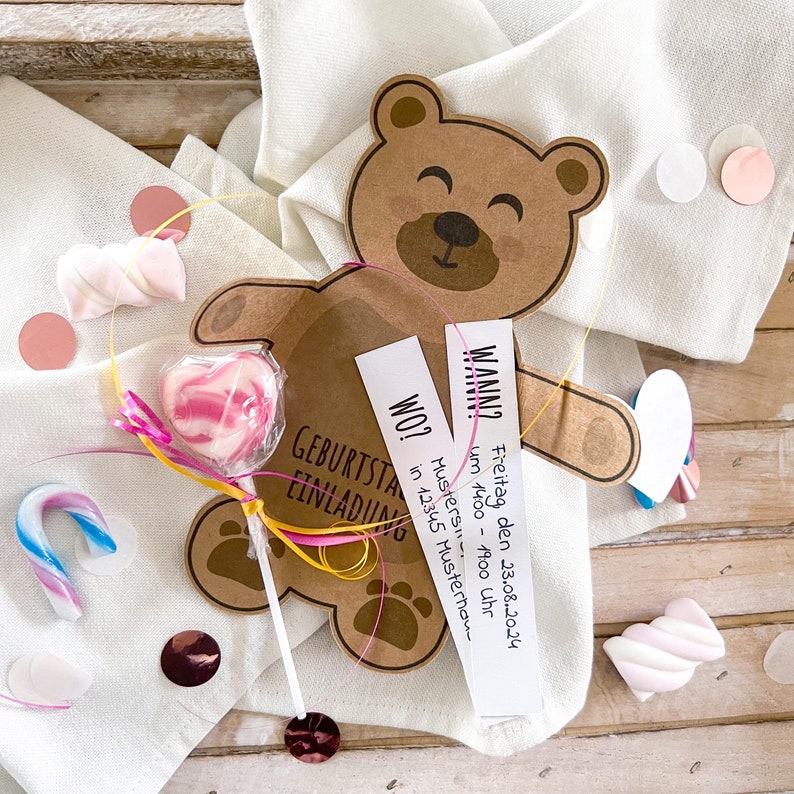 May include: A brown teddy bear-shaped invitation card with the text "GEBURTSTAG EINLADUNG". The card has two attached white labels with event details. A heart-shaped lollipop and candies are also present.