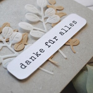 May include: A close-up of a handmade card with a light gray background. The card features white and tan paper cut-out leaves and a white rectangular label with the words "danke f&uuml;r alles" printed in black.