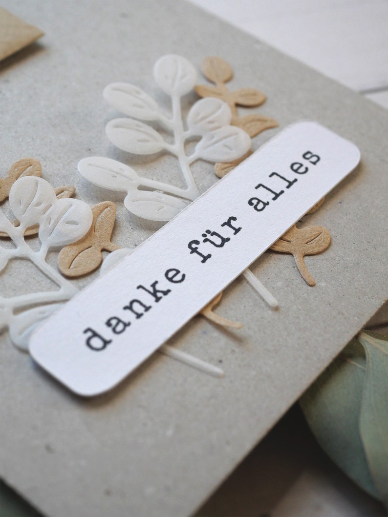 May include: A close-up of a handmade card with a light gray background. The card features white and tan paper cut-out leaves and a white rectangular label with the words "danke f&uuml;r alles" printed in black.