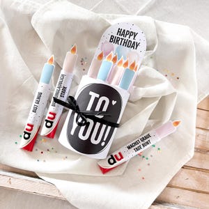 May include: A birthday gift set featuring candle-shaped cards with text in German. The set includes a card holder with "TO YOU!" and "HAPPY BIRTHDAY" text, tied with a black ribbon. The candles are white, blue, and pink with orange flames.