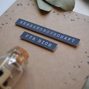 May include: A brown card with black text that reads "Herzensbotschaft f&uuml;r dich" and a small glass bottle with a cork stopper.