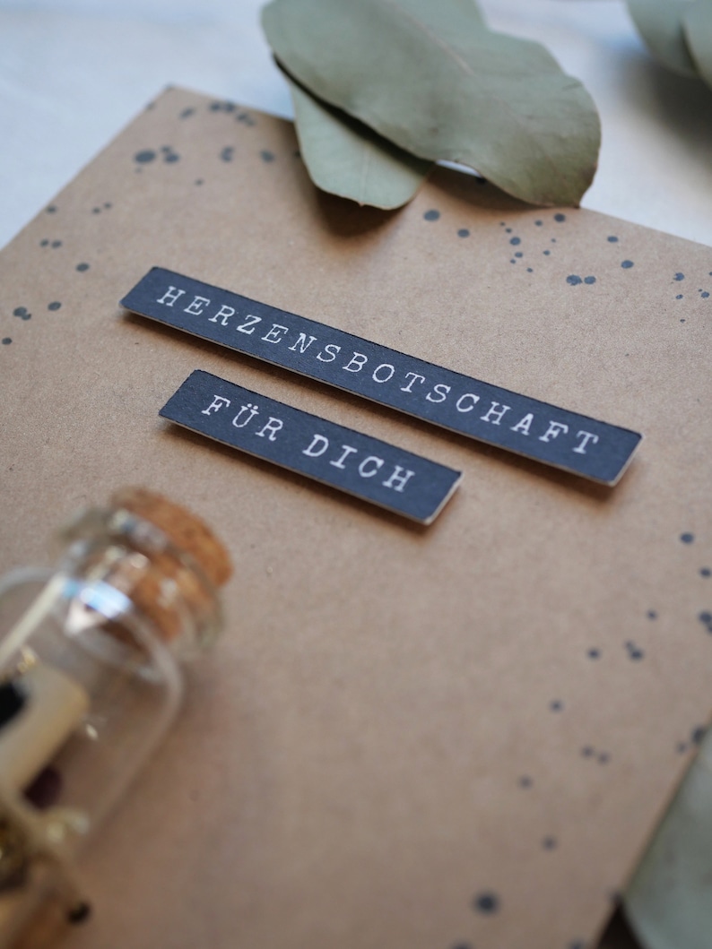 May include: A brown card with black text that reads "Herzensbotschaft f&uuml;r dich" and a small glass bottle with a cork stopper.