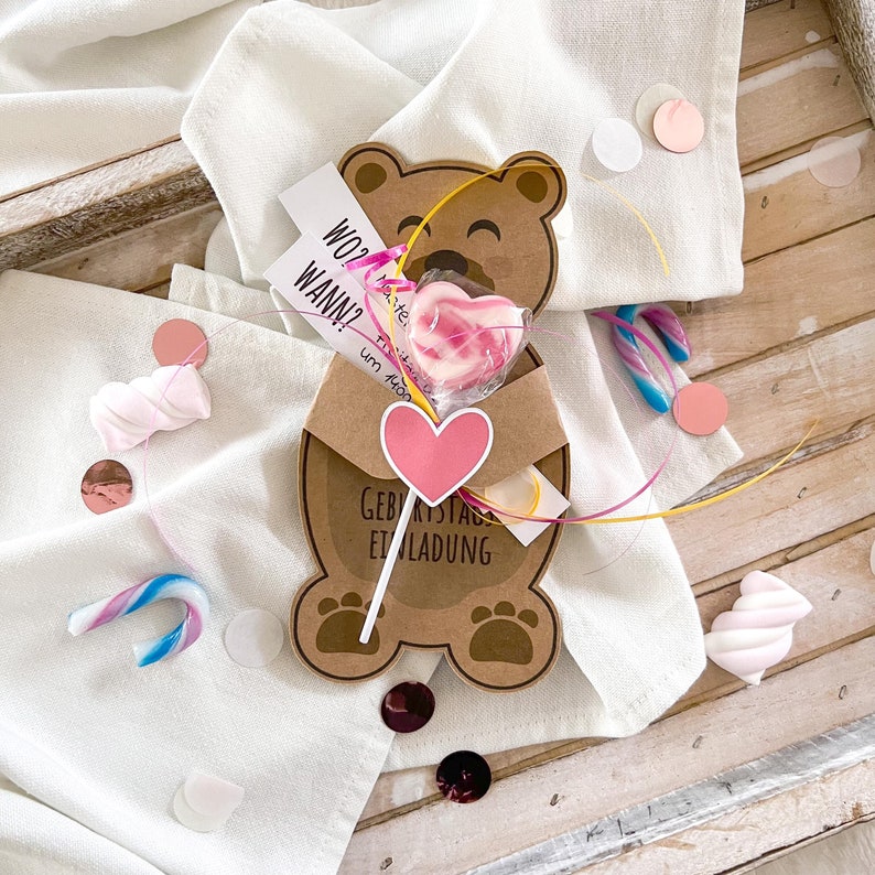 May include: A brown teddy bear-shaped invitation card with the text "Geburtstag Einladung" and a pink heart. A heart-shaped lollipop is attached. The card is on a white cloth with confetti and candies.