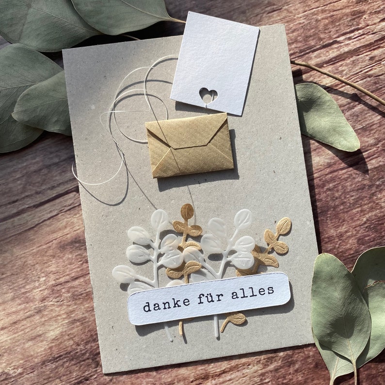 May include: A handmade greeting card with a kraft paper base, featuring a small kraft paper envelope, a white tag with a heart cutout, and decorative white and gold leaf accents. The card includes the text "danke f&uuml;r alles" on a white label.