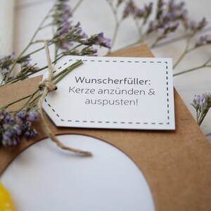 Puede incluir: Una tarjeta de papel marr&oacute;n con una etiqueta blanca adjunta. La etiqueta tiene el texto "Wunsch erf&uuml;ller: Kerze anz&uuml;nden & auspusten!" La tarjeta est&aacute; decorada con flores moradas secas.
