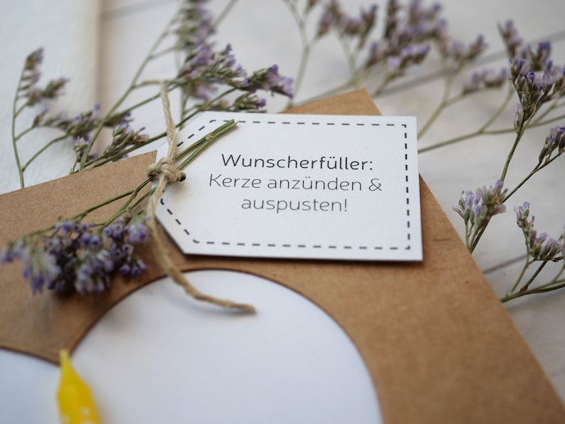 Puede incluir: Una tarjeta de papel marr&oacute;n con una etiqueta blanca adjunta. La etiqueta tiene el texto "Wunsch erf&uuml;ller: Kerze anz&uuml;nden & auspusten!" La tarjeta est&aacute; decorada con flores moradas secas.