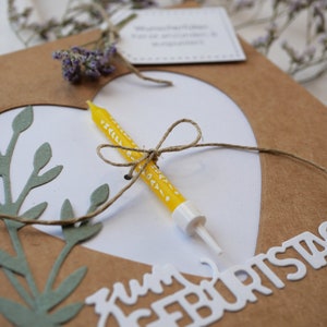 Puede incluir: Una tarjeta marr&oacute;n con un coraz&oacute;n blanco recortado. El coraz&oacute;n tiene una vela amarilla con lunares blancos atada con una cuerda. La tarjeta dice "Zum Geburtstag" en letras blancas.