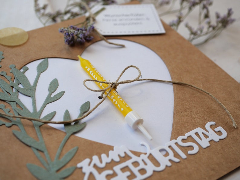 Puede incluir: Una tarjeta marr&oacute;n con un coraz&oacute;n blanco recortado. El coraz&oacute;n tiene una vela amarilla con lunares blancos atada con una cuerda. La tarjeta dice "Zum Geburtstag" en letras blancas.