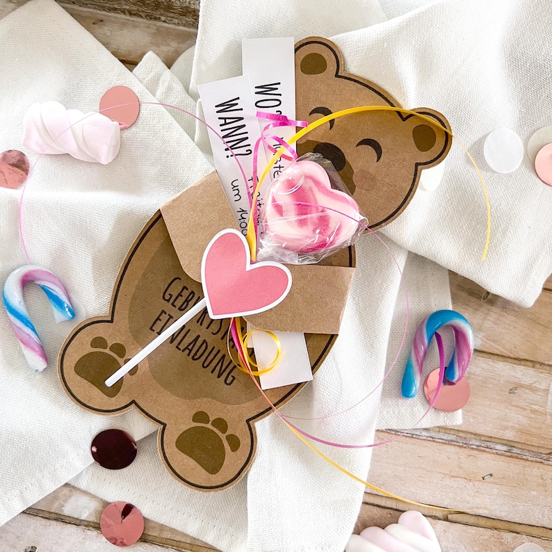 May include: A brown bear-shaped invitation card with a pink heart-shaped lollipop and candy canes. The card has text in German and is decorated with pink and yellow ribbons and confetti.