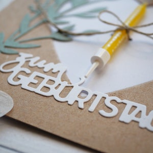 Puede incluir: Una tarjeta marr&oacute;n con las palabras "Feliz cumplea&ntilde;os" escritas en letras blancas. Una vela amarilla con una cuerda atada alrededor est&aacute; en la tarjeta.