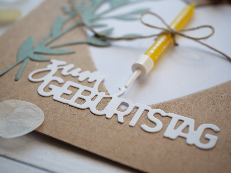 Puede incluir: Una tarjeta marr&oacute;n con las palabras "Feliz cumplea&ntilde;os" escritas en letras blancas. Una vela amarilla con una cuerda atada alrededor est&aacute; en la tarjeta.