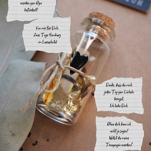 May include: A small glass bottle with a cork top is tied with twine and a black heart charm. The bottle contains a rolled up piece of paper and gold leaf. The bottle is sitting on a brown surface with a piece of paper that says "Danke, dass du mich jeden Tag zum L&auml;cheln bringst. Ich liebe Dich." and another piece of paper that says "Ohne dich kann ich nicht ja sagen! Willst du meine Trauzeugin werden?"