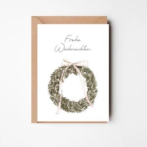 Könnte beinhalten: Eine Weihnachtskarte mit einem Aquarellkranz und einer hellrosa Schleife. Auf der Karte steht in zarter Schrift „Frohe Weihnachten“. Die Karte befindet sich auf einem braunen Umschlag.