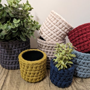 May include: A set of six crocheted plant pots in various colors, including gray, beige, red, blue, and yellow. The pots are made with a chunky yarn and have a textured, woven look. A small green plant is in one of the pots.