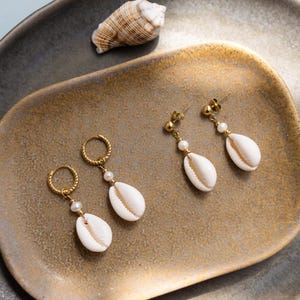 Könnte beinhalten: Goldfarbene Ohrringe mit weißen Muschelanhängern und kleinen Perlenakzenten. Die Ohrringe werden auf einem goldfarbenen Tablett mit einer Muschel präsentiert. Die Ohrringe haben ein Stecker- und Creolen-Design.