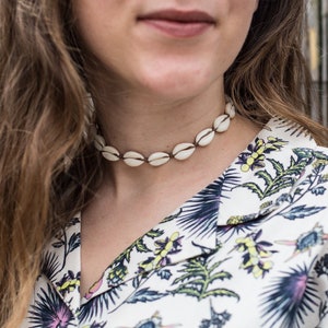 Può includere: Una donna con lunghi capelli castani indossa una camicia bianca con un motivo floreale e una collana di conchiglie bianche.