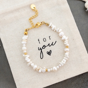 Könnte beinhalten: Ein Armband aus weißen Perlen und goldenen Perlen mit einem Verschluss. Das Armband wird auf einem weißen Stoffbeutel mit den Worten "for you" in Schwarz präsentiert.