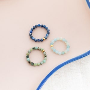 Könnte beinhalten: Drei Perlenringe auf einer rosa Oberfläche. Die Ringe bestehen aus blauen, grünen und hellblauen Perlen. Jeder Ring hat einen goldenen Perlenakzent.
