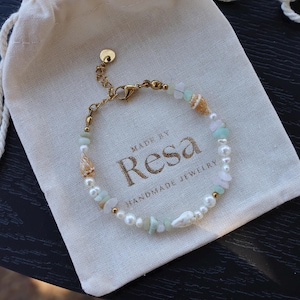 Könnte beinhalten: Ein handgefertigtes Armband mit Perlen, Muscheln und hellgrünen Steinen. Das Armband liegt auf einem cremefarbenen Kordelzugbeutel mit dem Text "MADE BY Resa HANDMADE JEWELRY". Das Armband hat eine goldfarbene Kette und einen Verschluss.