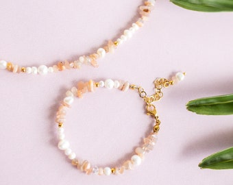 Könnte beinhalten: Eine zarte goldene Kettenkette und ein Armband mit abwechselnden Sunstone- und Perlenperlen. Der Schmuck wird auf einem rosa Hintergrund mit grünen Blättern präsentiert.