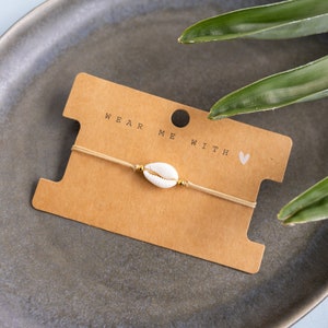 Könnte beinhalten: Ein schlichtes Armband mit einem weißen Muschel-Anhänger und goldenen Perlen auf einer beigefarbenen Schnur. Das Armband wird auf einer braunen Karte mit dem Text "Wear Me With" und einem kleinen Herz präsentiert.