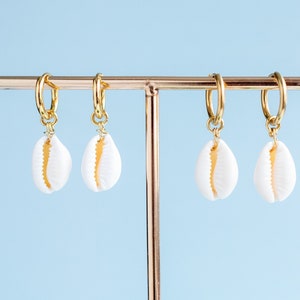 May include: Four gold hoop earrings with white cowrie shell charms hanging from them. The earrings are displayed on a gold stand against a light blue background.