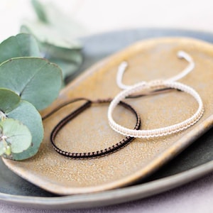 Op de afbeelding: Twee kralenarmbanden op een klein, lichtbruin keramiek schaaltje. Het schaaltje ligt op een groter, grijs keramiek schaaltje. Een takje eucalyptusbladeren bevindt zich aan de linkerkant van de afbeelding.
